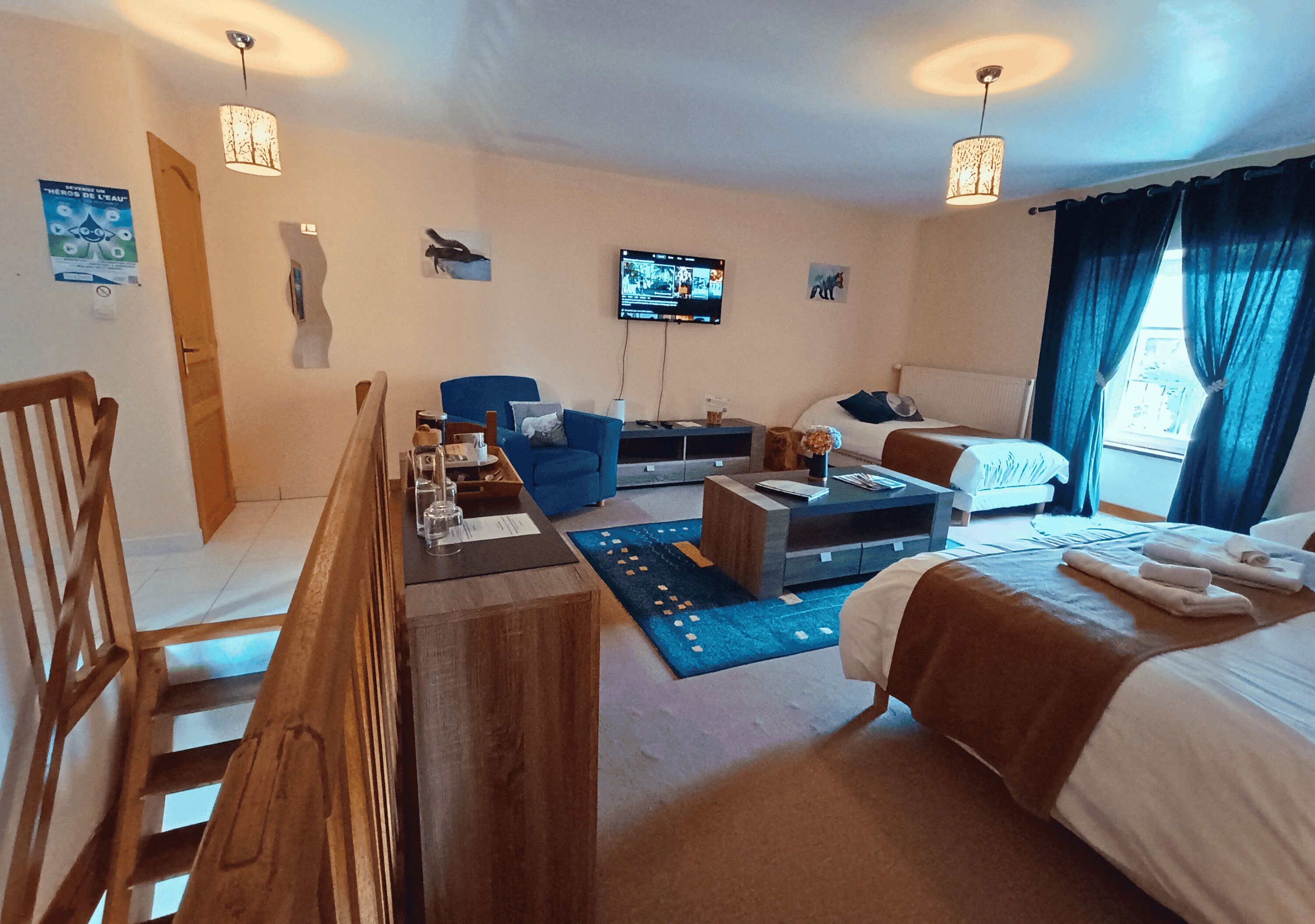 A hotel room with a wooden staircase, a blue sofa, a bed with a brown blanket, a flat-screen TV, and a window with curtains.
