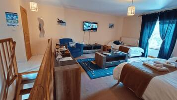 A hotel room with a wooden staircase, a blue sofa, a bed with a brown blanket, a flat-screen TV, and a window with curtains.