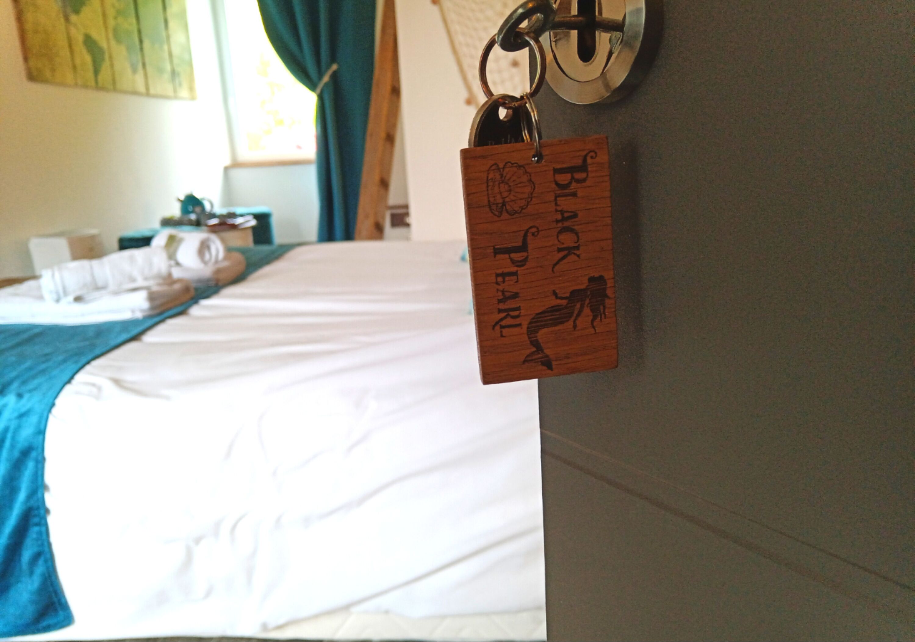 Una cama bien hecha con una manta azul y un llavero de madera con la inscripción 'BLACK PEARL' colgado en la pared.