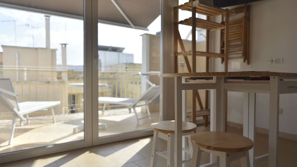 Ein moderner Raum mit Glastür zu einem Balkon mit Sonnenliegen. Zwei Holzschemel und ein kleiner Tisch sind vorhanden.
