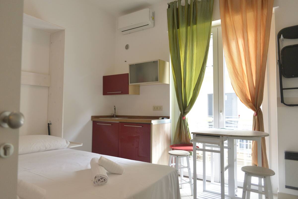 Hotellihuone, jossa on sänky, pieni keittokomero, ikkuna verhoilla ja parveke.