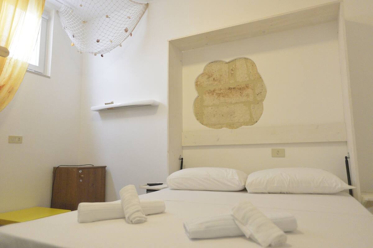 Hotellihuone, jossa on sänky, valkoiset vuodevaatteet, puinen kaappi, pieni pöytä ja seinälle asennettu ilmastointilaite.