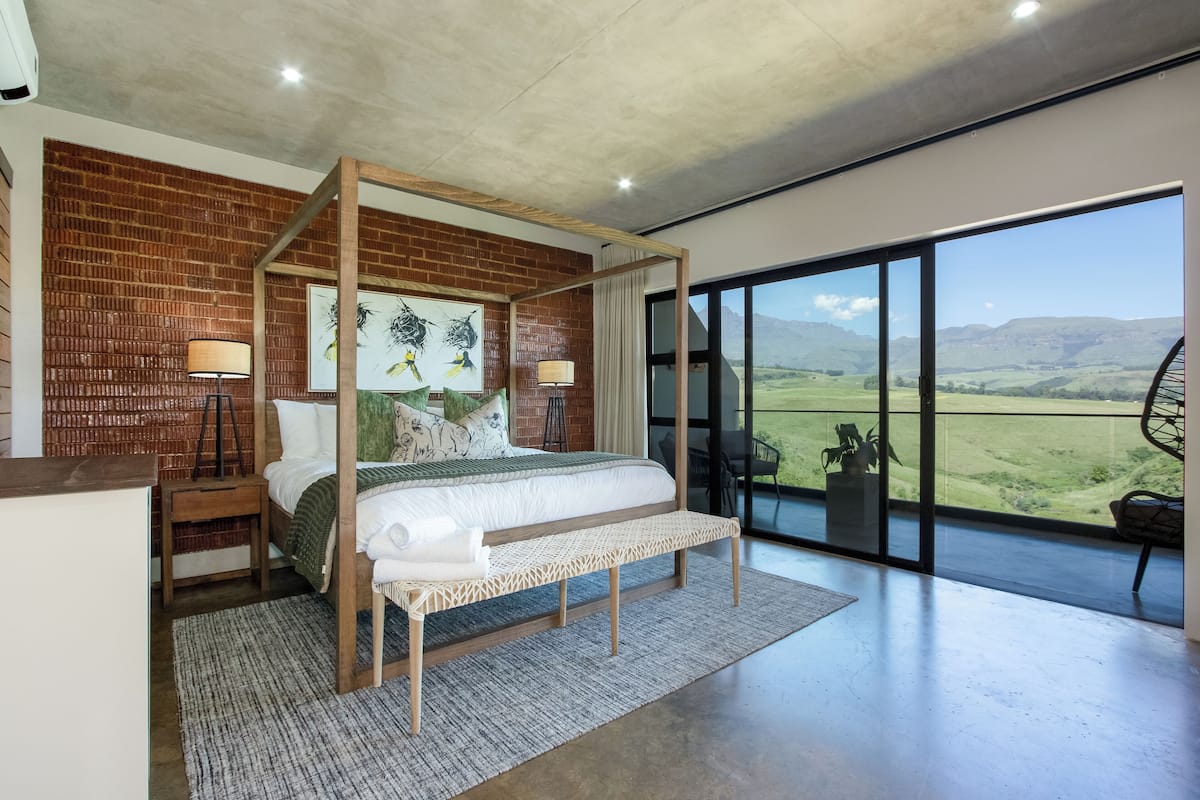 A modern bedroom with a four-poster bed, a large window with a view of the countryside, and a balcony with outdoor seating.