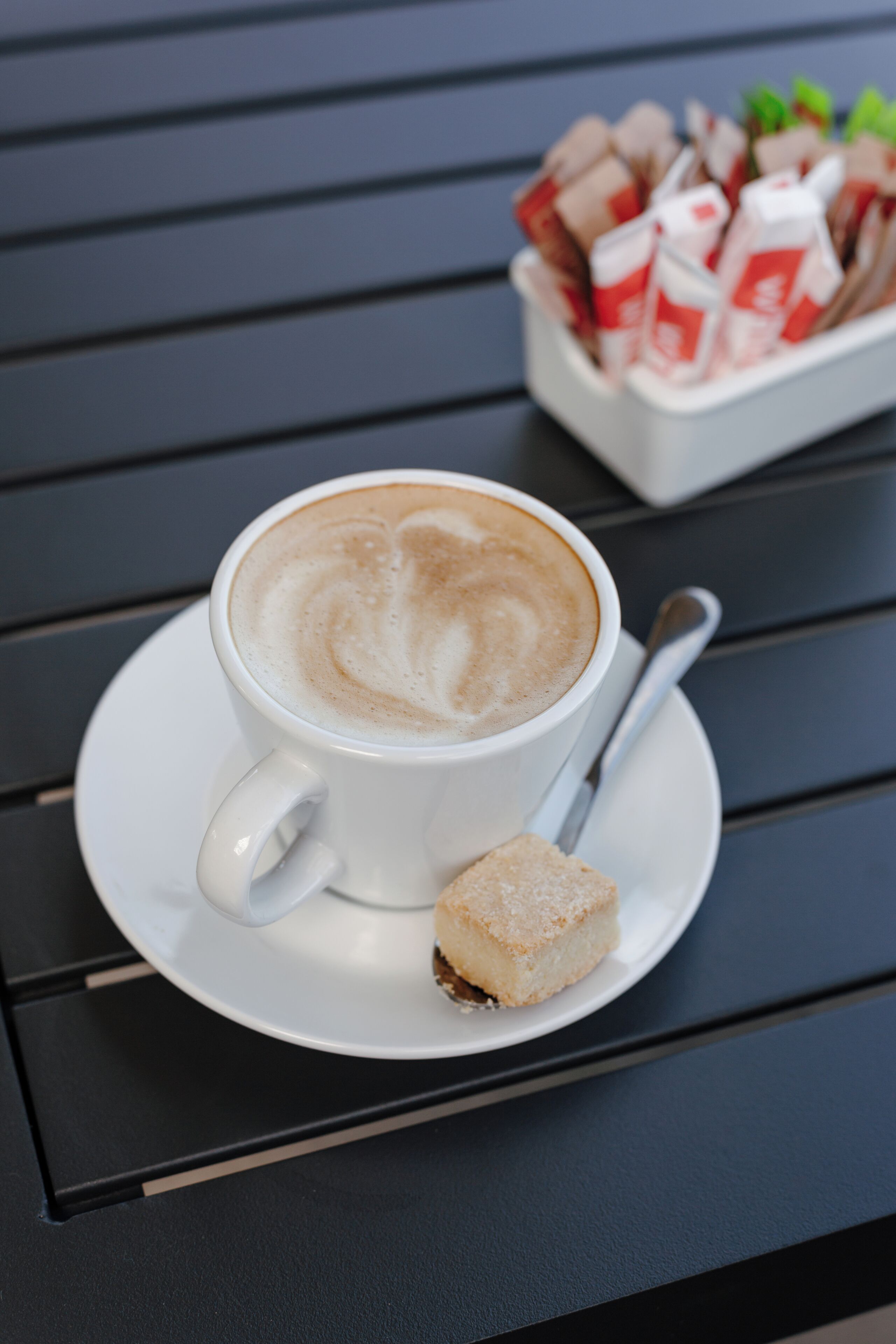 Eine Tasse Kaffee mit Latte-Art auf einer weißen Untertasse, dazu ein kleines Stück Kuchen und eine Biscotti-Verpackung auf einem dunklen Tisch.