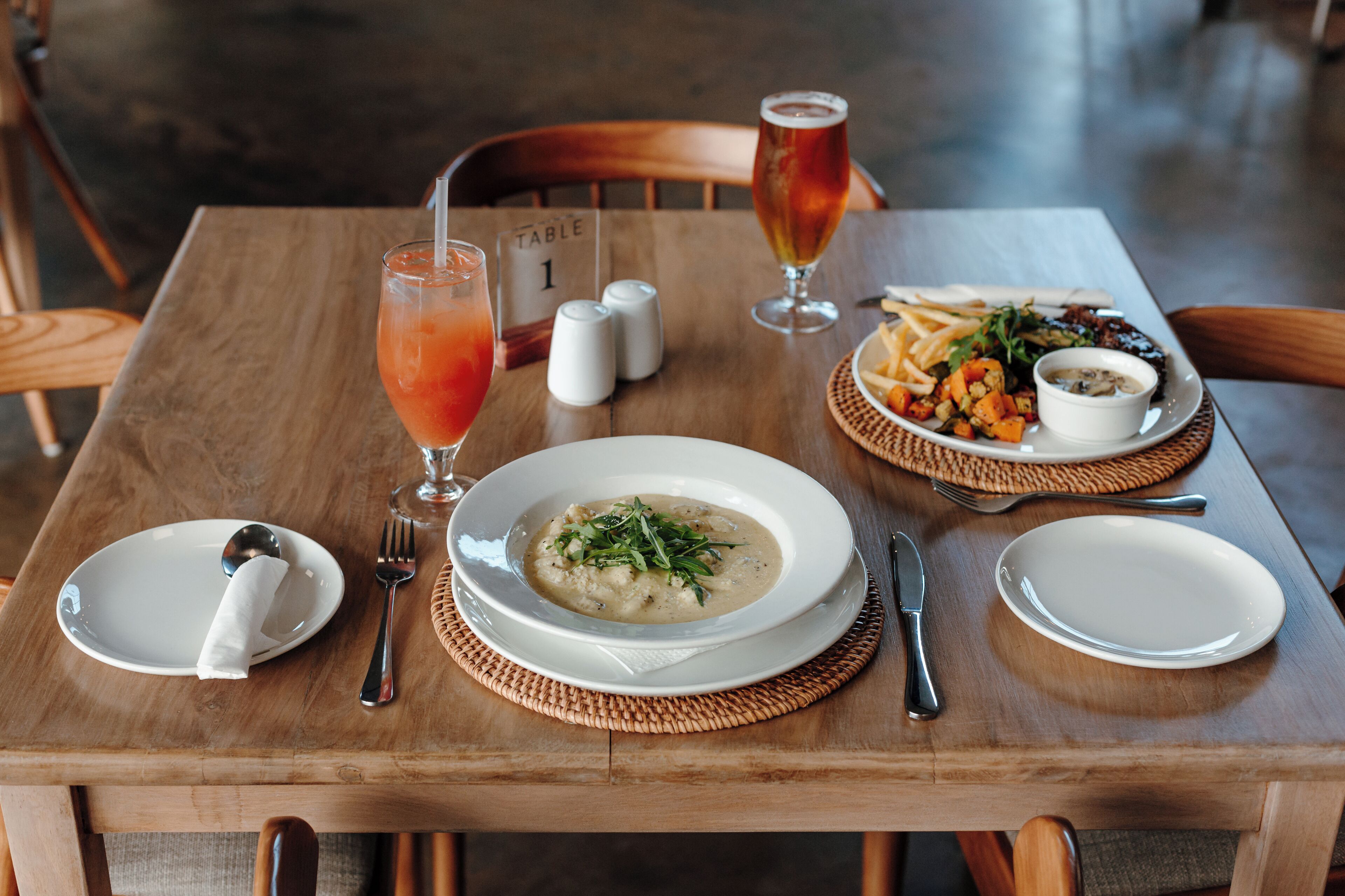 Una mesa de madera con un tazón de sopa, un vaso de bebida roja, un plato de verduras y un recipiente pequeño de salsa.