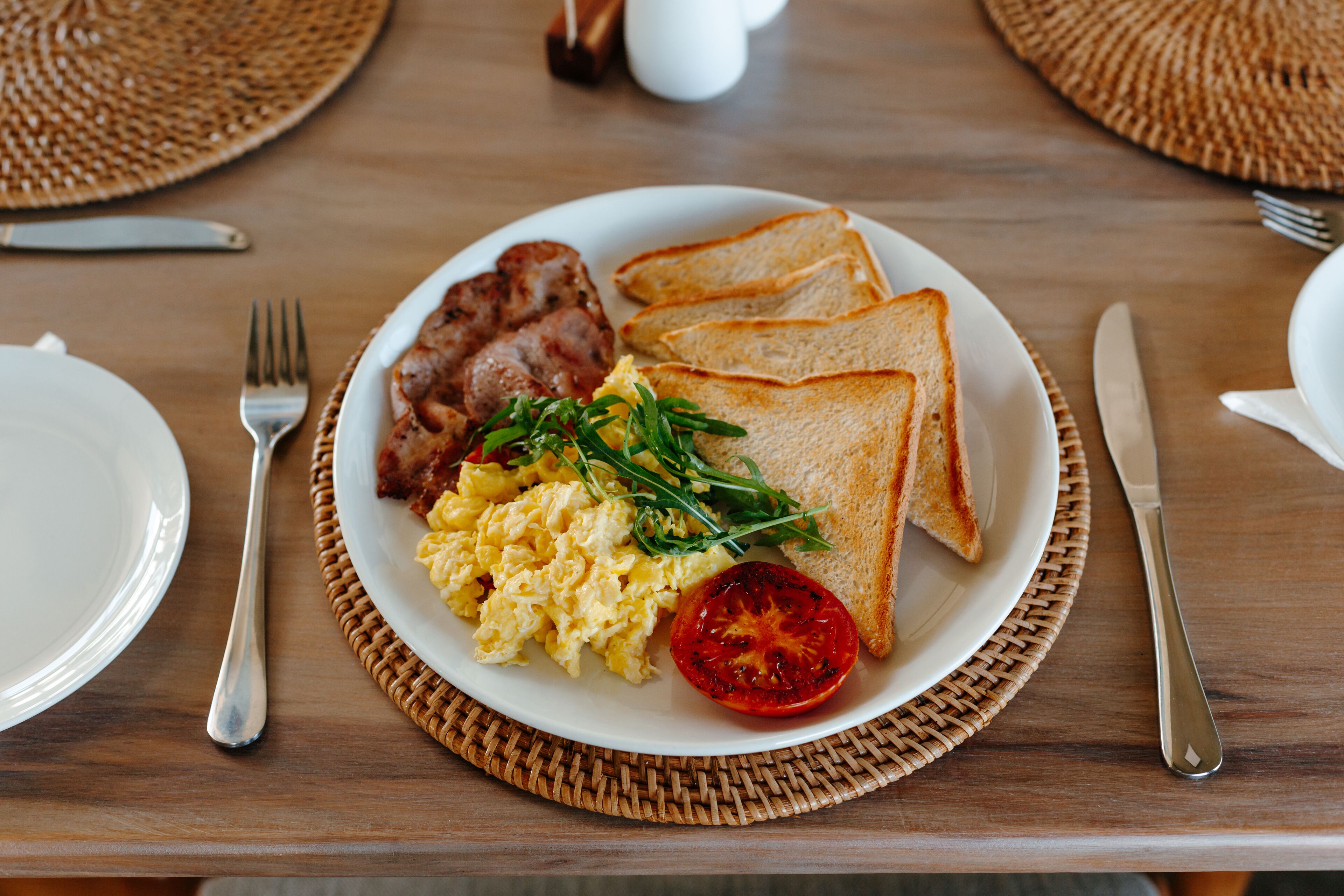 A breakfast plate with scrambled eggs, bacon, toast, and a tomato slice.