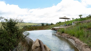 A natural water feature with rocks and greenery, a shaded area with a table and chairs, and a view of distant hills. - Dragonview Lodge (Cathkin Park)