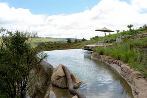 Un elemento acuático natural con rocas y vegetación, una zona sombreada con una mesa y sillas, y vistas a colinas distantes.