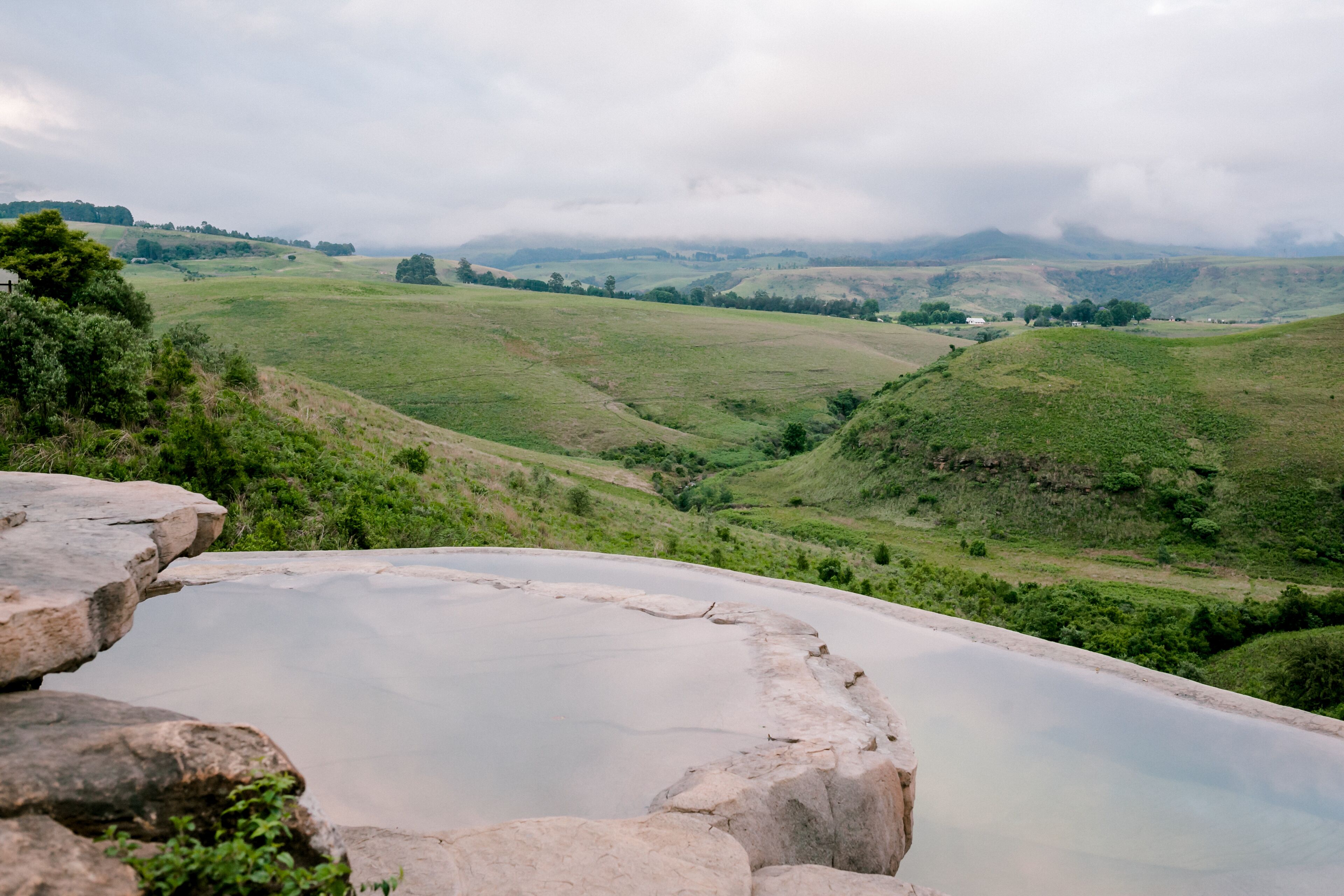Une vue panoramique d’une vallée avec une vaste étendue d’eau calme, entourée de collines verdoyantes et surplombée par un ciel nuageux.