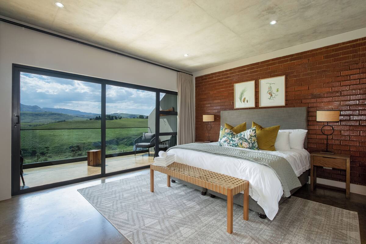 A bedroom with a large bed, a wooden bench, a nightstand, and a view of a green landscape through a sliding door.