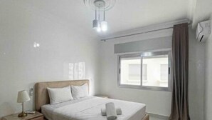 A modern bedroom with a large bed, bedside tables, a nightstand, a window with curtains, and a ceiling light fixture.