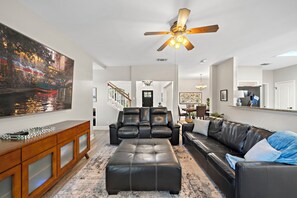 A living room with a large flat-screen TV, leather sofas, a wooden console, and a ceiling fan.