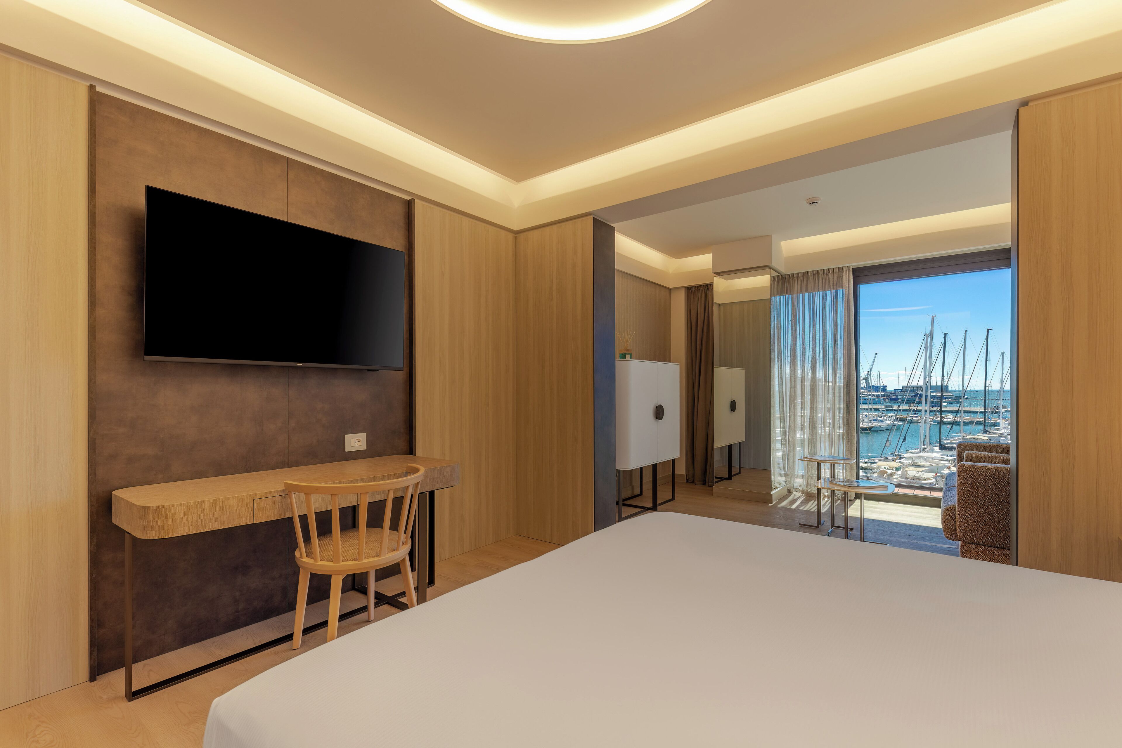 A modern hotel room with a large bed, a desk with a chair, a flat-screen TV mounted on the wall, and a view of a marina through a large window.