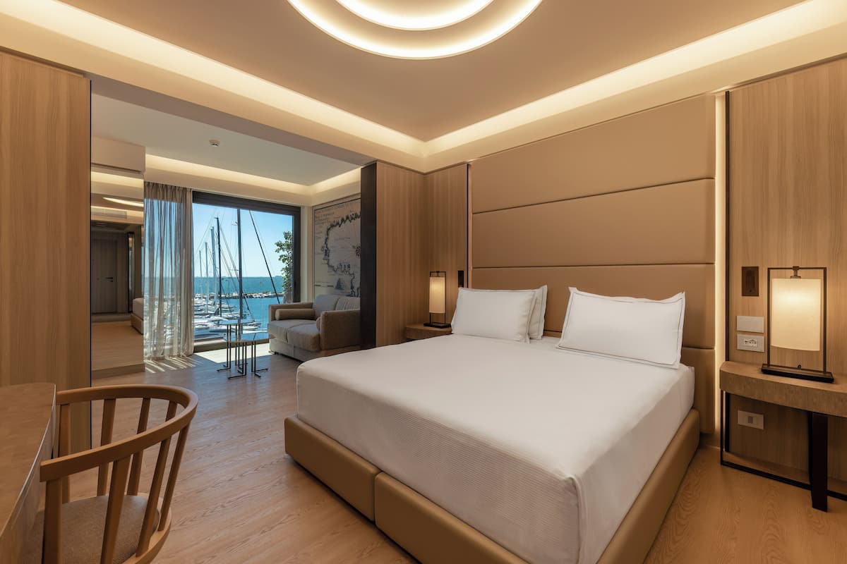 A modern hotel room with a large bed, a wooden chair, a desk, and a view of the sea.