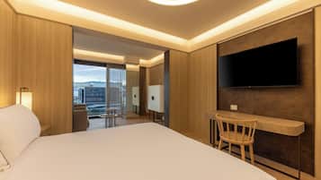 Une chambre d’hôtel moderne dotée d’un grand lit, d’un bureau avec une chaise, d’un téléviseur à écran plat fixé au mur et offrant une vue sur la ville grâce à de grandes fenêtres.