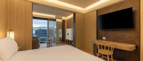 Una habitación de hotel moderna con una cama grande, un escritorio con silla, un televisor de pantalla plana montado en la pared y vistas a la ciudad a través de ventanas amplias.
