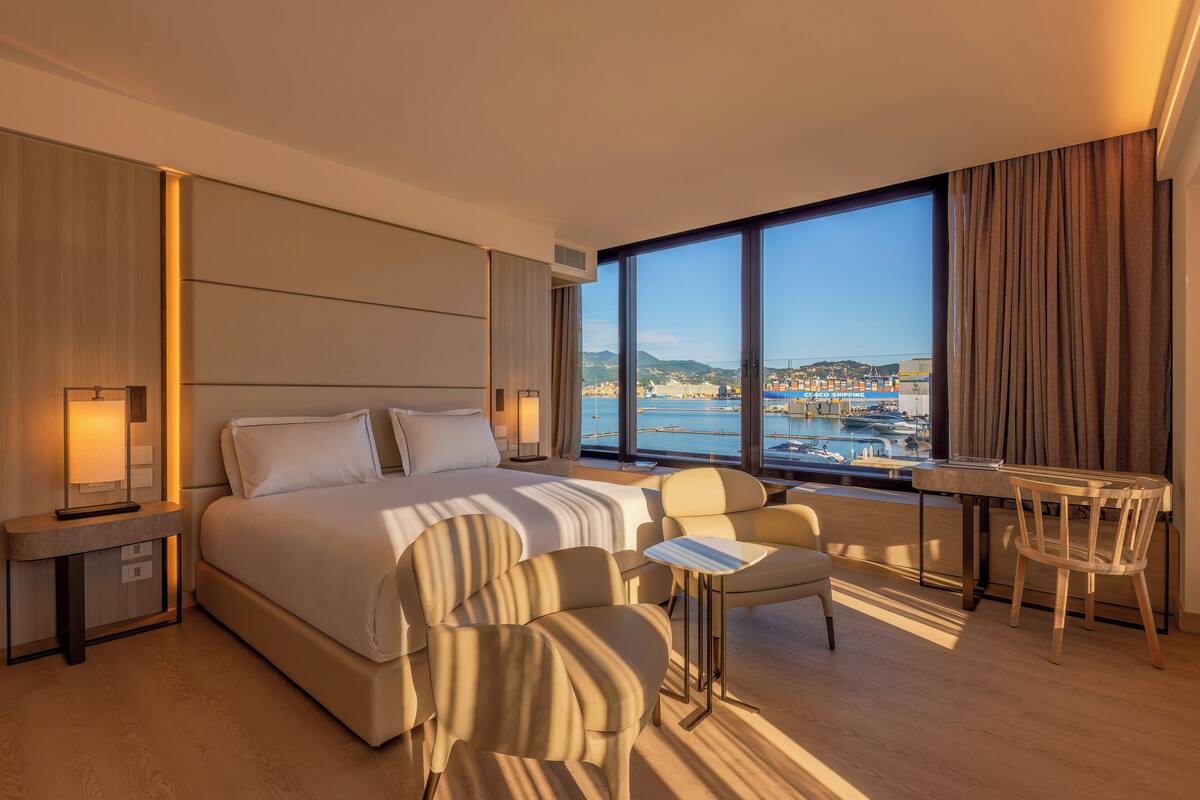 A modern hotel room with a large bed, a sitting area with chairs, a small table, and a view of a marina.