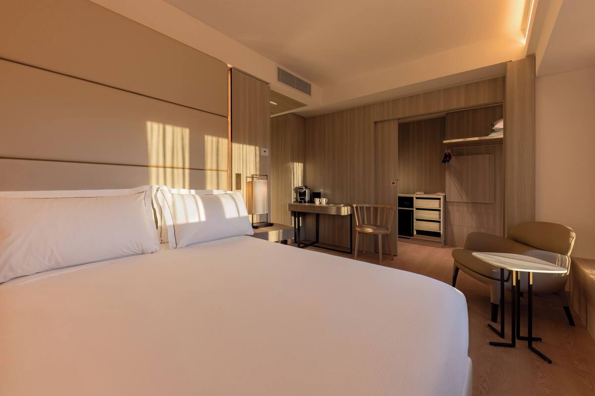 A hotel room with a large bed, a desk with a coffee maker, a chair, and a wardrobe.
