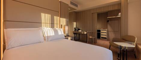 A hotel room with a large bed, a desk with a coffee maker, a chair, and a wardrobe.
