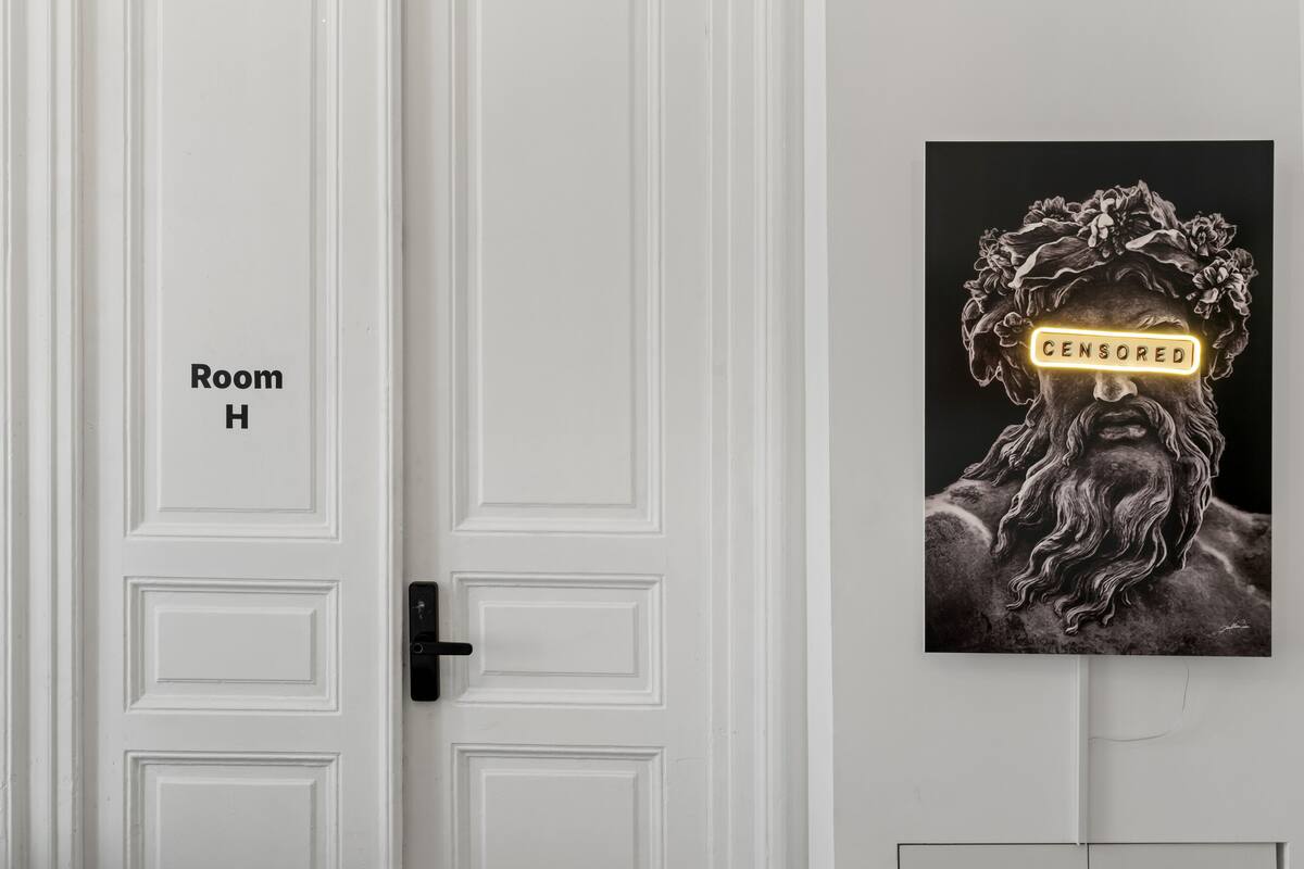 A white door labeled "Room H" with a black handle. Adjacent to the door is a framed artwork depicting a figure with a blurred face and the word "CENSORED" in a neon sign.