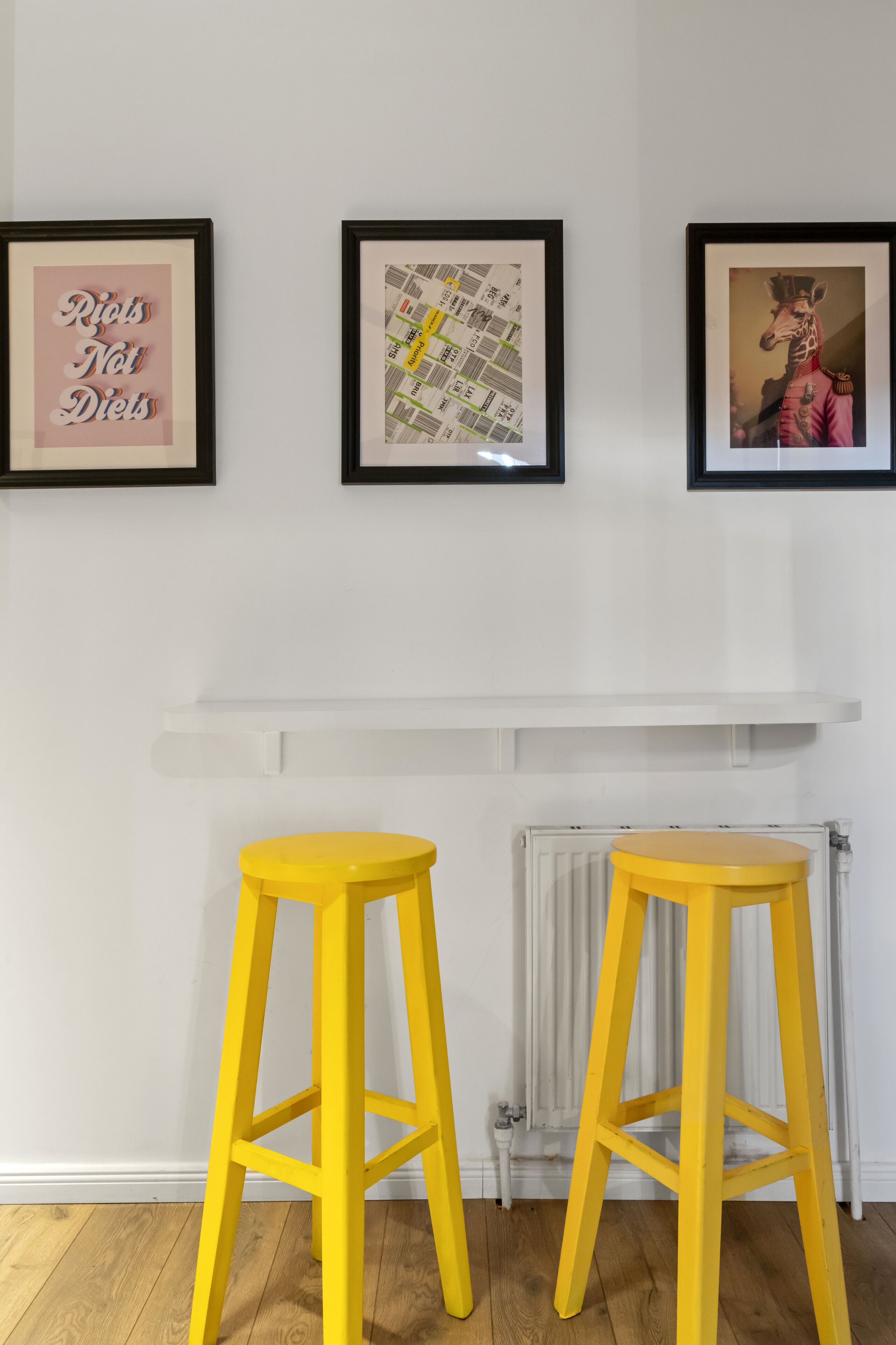 A room with two yellow stools, a white shelf, and three framed artworks on the wall.