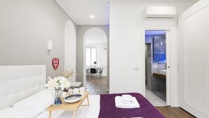 Ein modernes Hotelzimmer mit einem Bett, einem kleinen Tisch mit einer Vase und einer Schale, einer violetten Badmatte und Blick auf den Speisesaal.