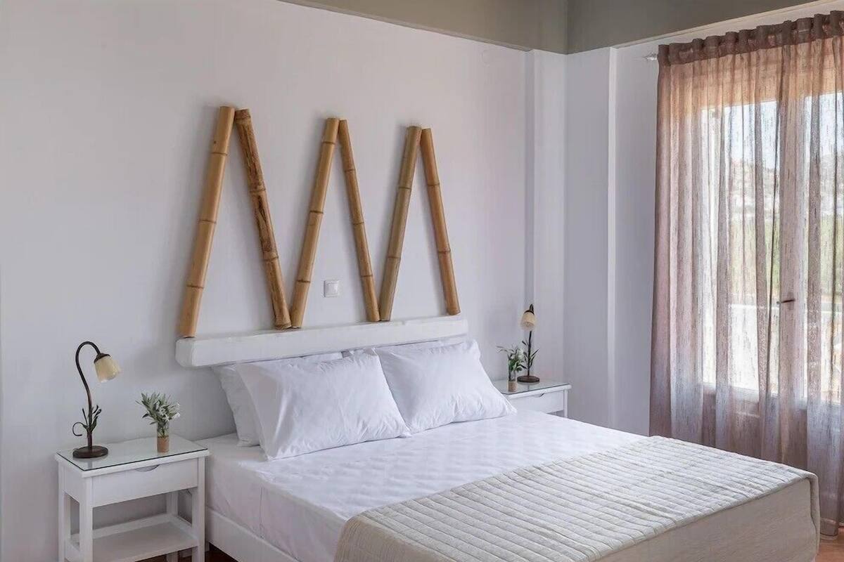 A neatly arranged bedroom with a large bed, white bedding, two bedside tables with lamps, and a window with sheer curtains.