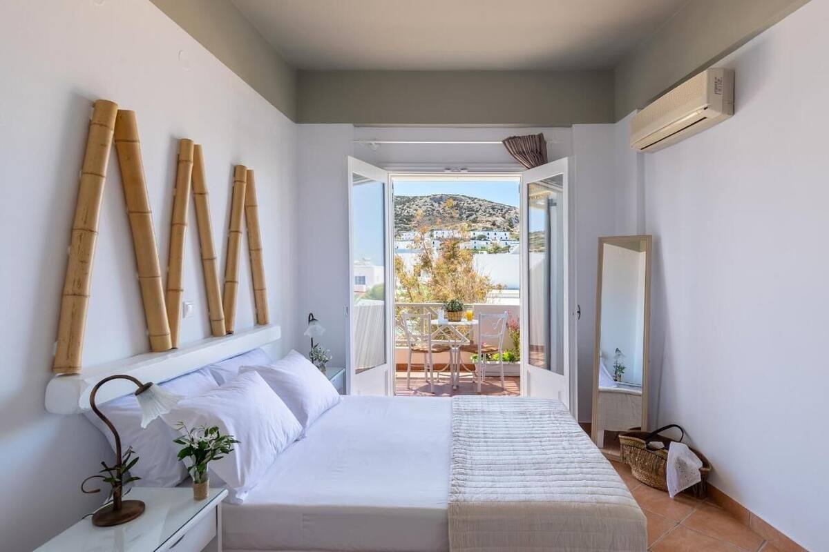 A bedroom with a bed, bedside table, lamp, and a view of a balcony with a table and chairs.