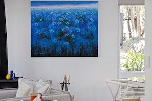 Una habitación con un cuadro grande de flores azules en la pared, un sofá gris, una mesita con una silla blanca y una ventana con vistas a zonas verdes.