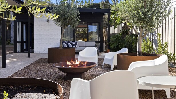 A modern outdoor seating area with a fire pit, white chairs, and a white building in the background.