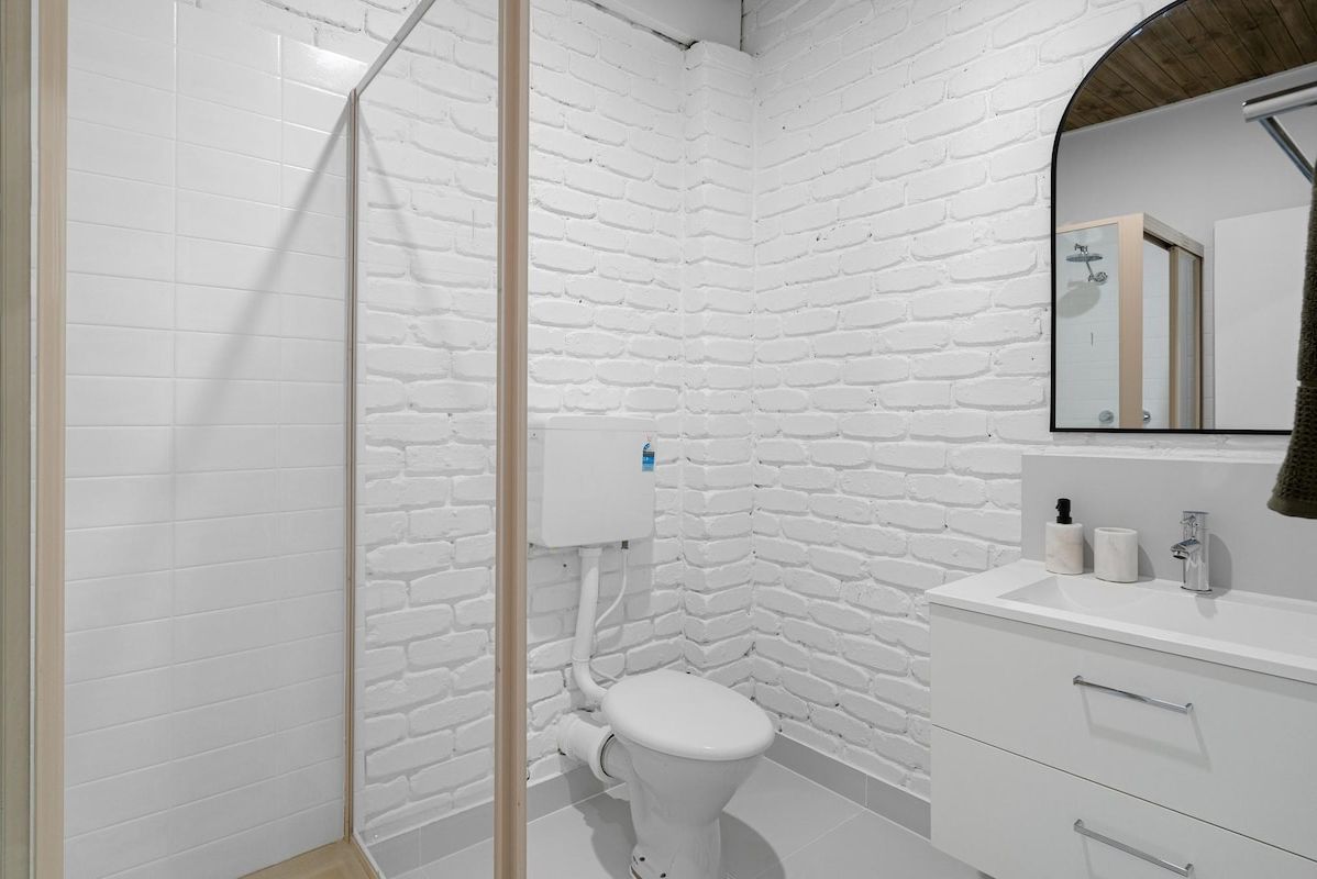 A bathroom with a white brick wall, a white toilet, a white sink with a mirror, and a shower area.