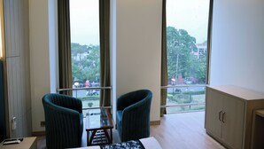 A room with two chairs, a small table, and a cabinet. Large windows offer a view of greenery and buildings outside. - Hotel Casa Royal (Amritsar)
