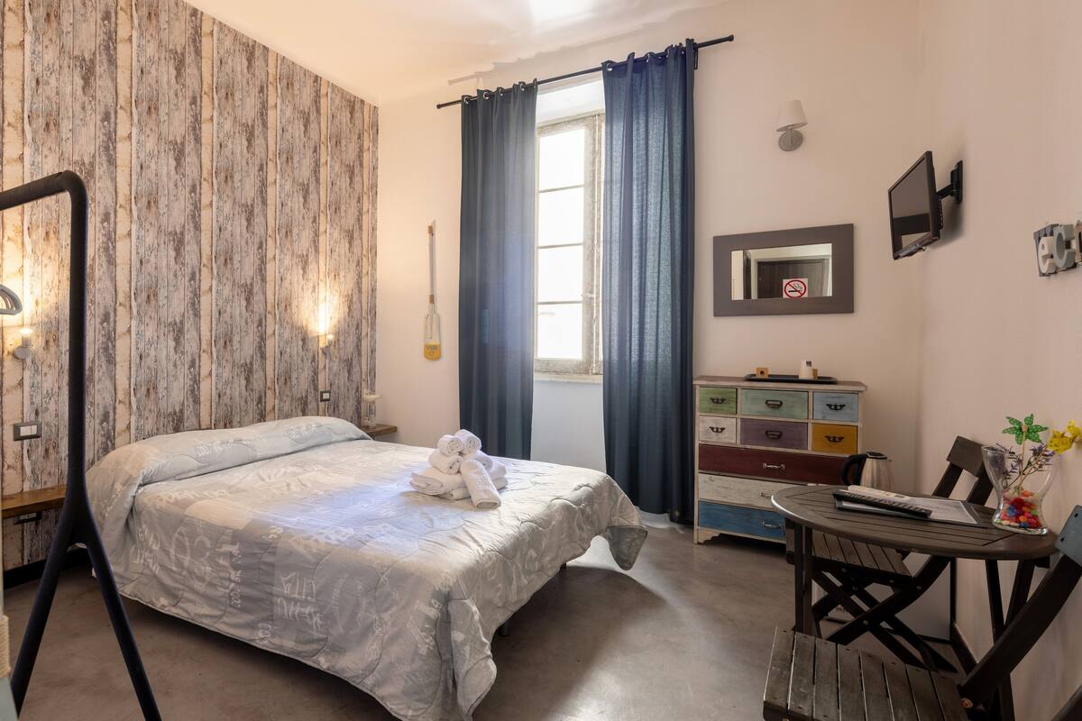Une chambre à coucher comprenant un lit, une commode, un bureau avec une chaise, une fenêtre avec des rideaux, un miroir et un téléviseur fixé au mur.