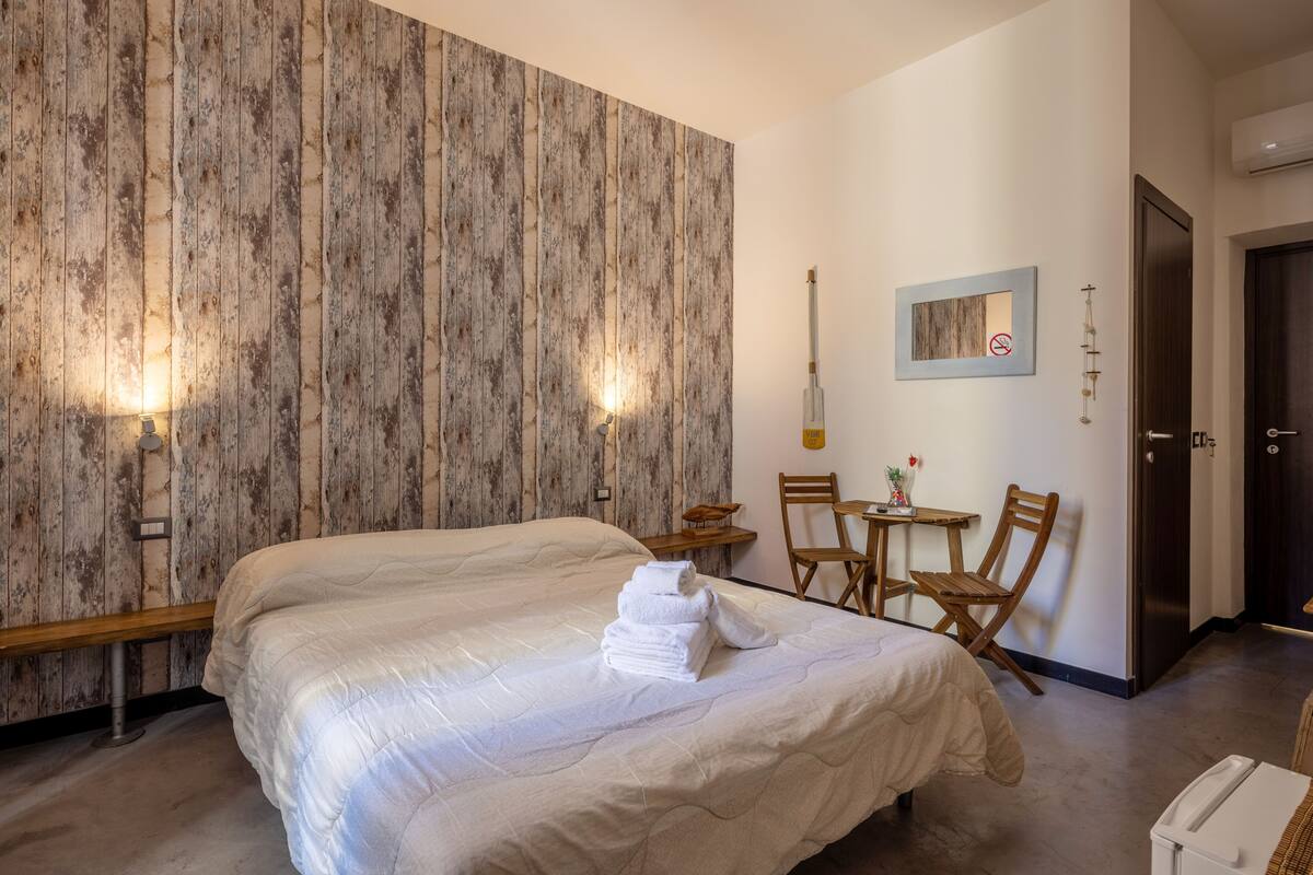 Une chambre avec un grand lit, un lambris en bois, une petite table avec des chaises, un luminaire fixé au mur et une porte.