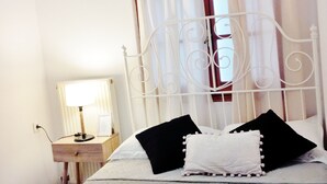 Una cama bien hecha con cabecera blanca, una mesita de noche con lámpara y una ventana con cortinas.