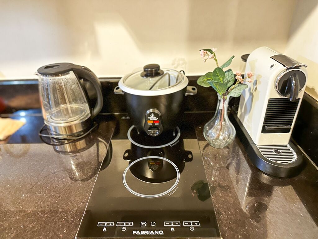 Una cocina con una placa de inducción negra, un hervidor de vidrio, una olla negra con tapa y un hervidor eléctrico blanco.