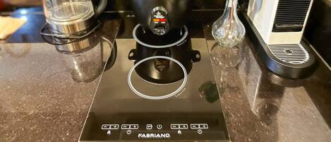 Una cocina con una placa de inducción negra, un hervidor de vidrio, una olla negra con tapa y un hervidor eléctrico blanco.
