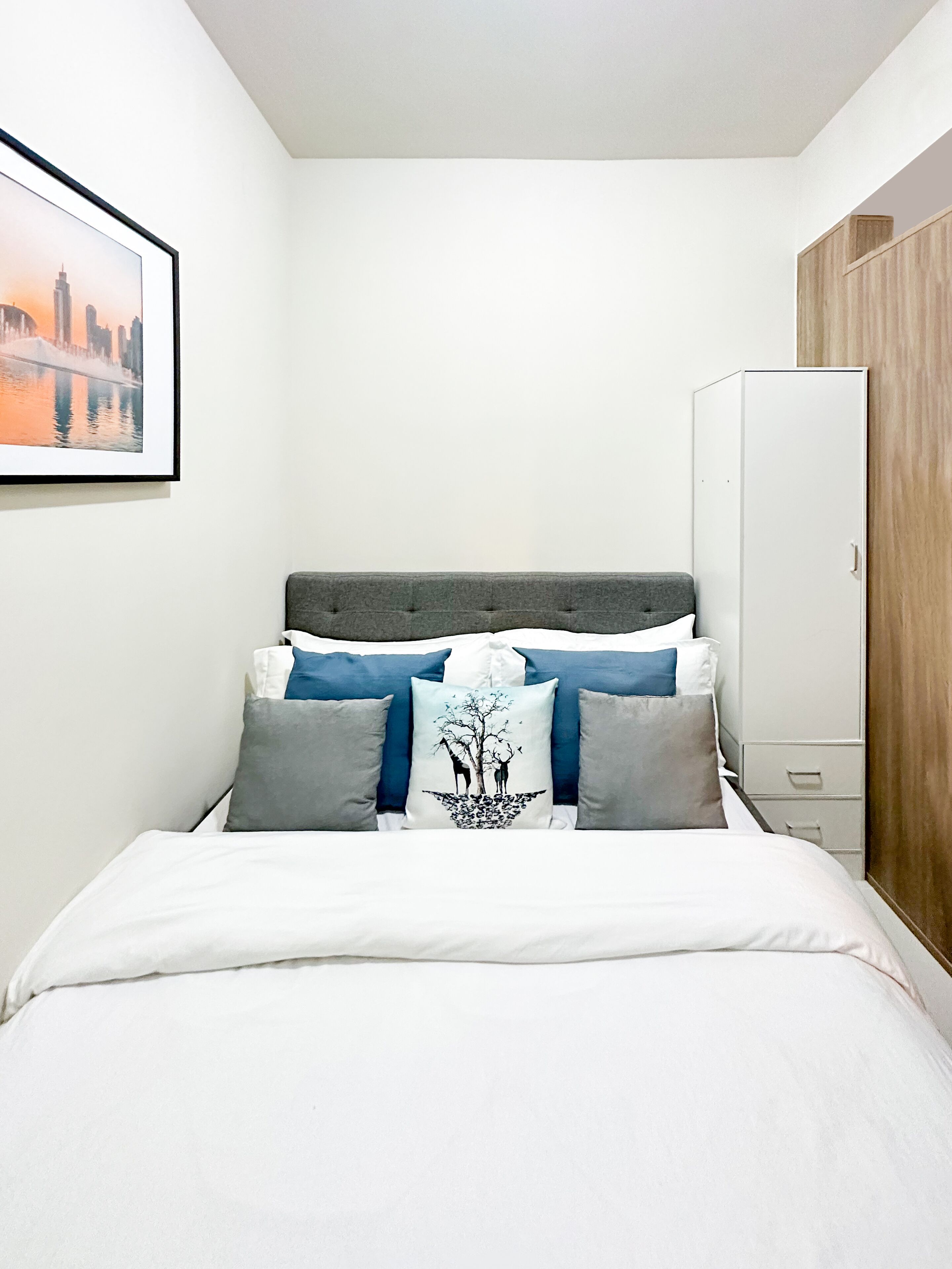 A neatly made bed with a grey headboard, a framed picture on the wall, and a wooden wardrobe.