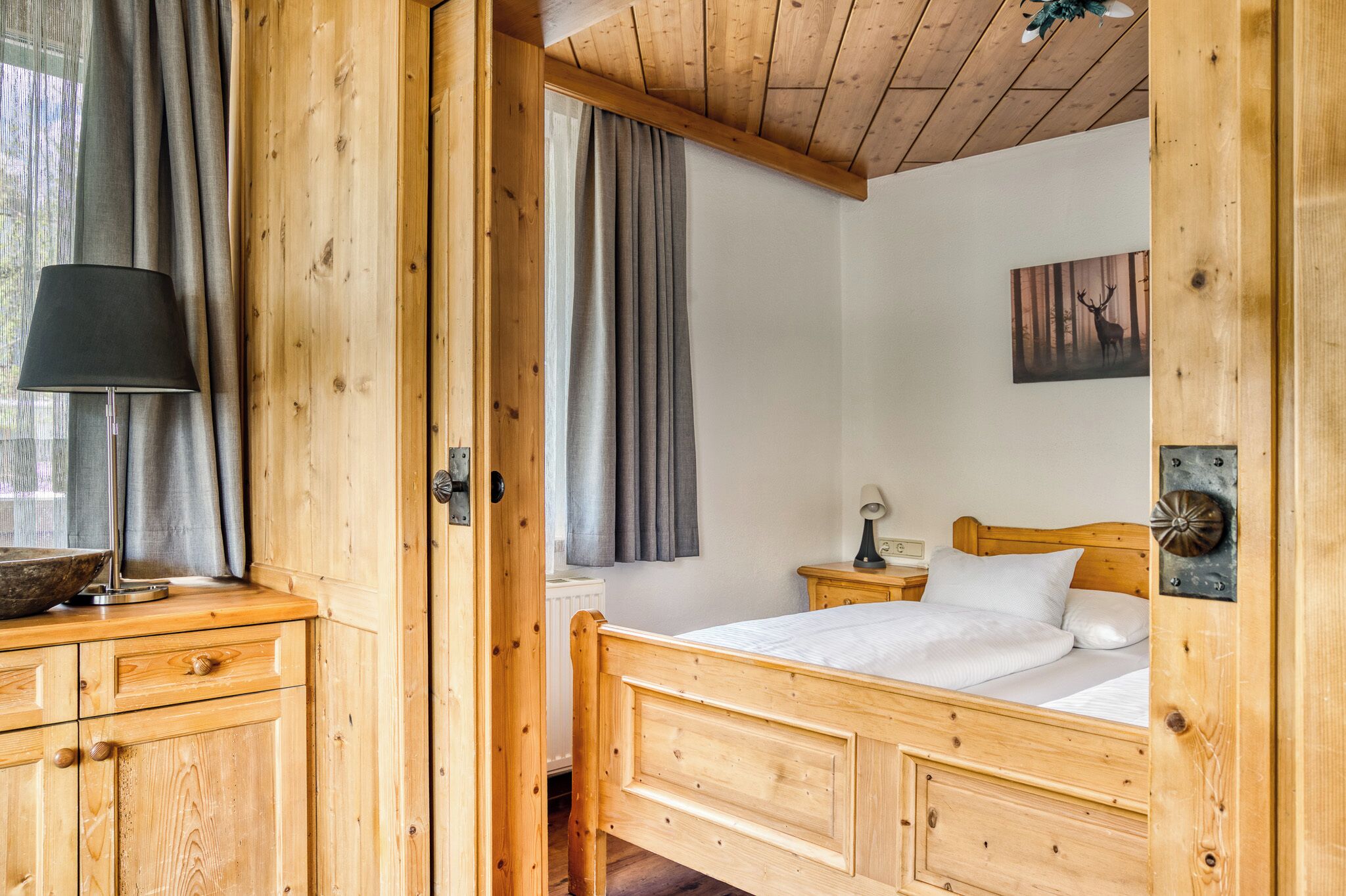 A wooden room with a bed, a nightstand, a lamp, and a window with curtains.