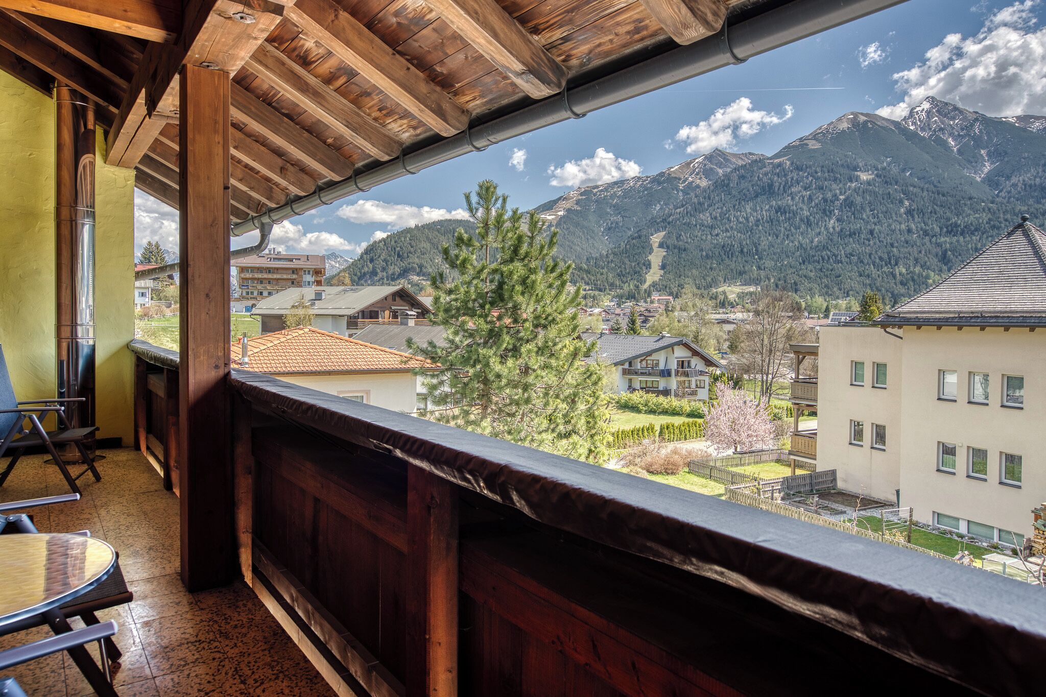 A balcony with a wooden ceiling overlooking a mountain village with houses and a ski slope.