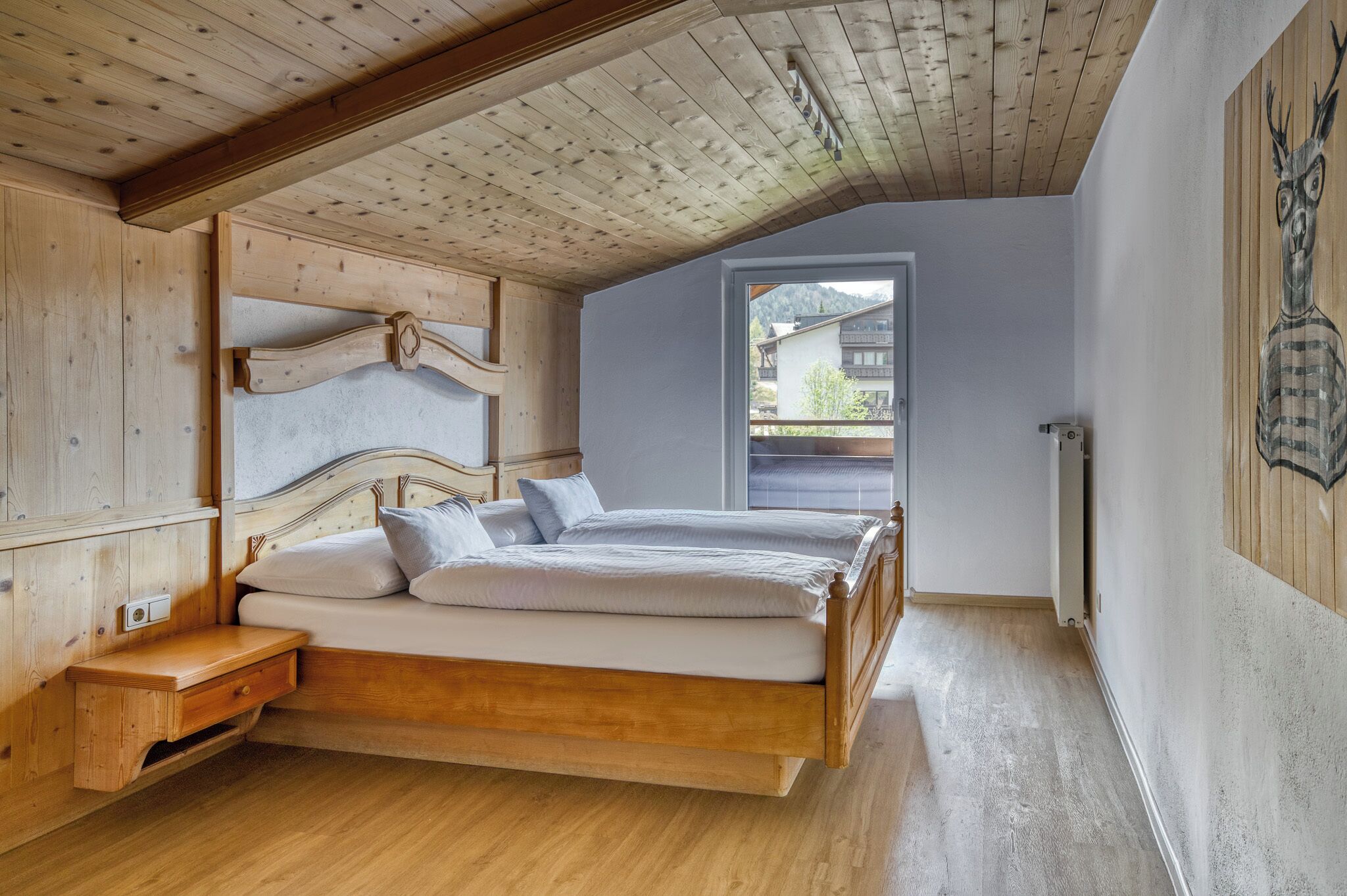 A bedroom with a sloped ceiling, wooden headboard, two beds, a nightstand, and a window with a view of buildings outside.
