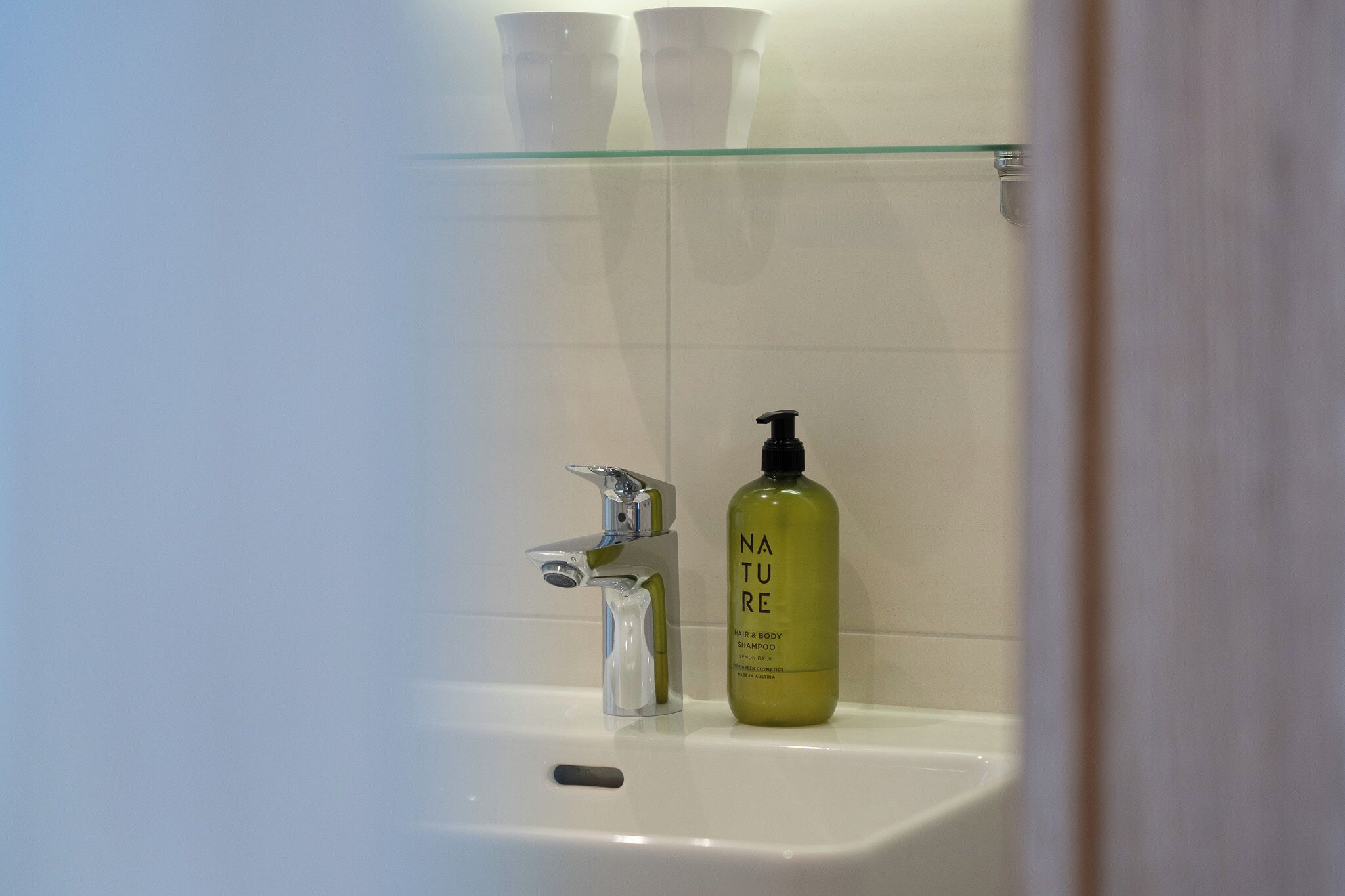 Baño con repisa de vidrio que sostiene tres tazas blancas, una botella de vidrio transparente de champú y un grifo moderno.