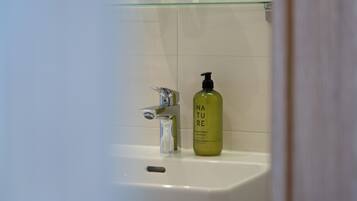 Baño con repisa de vidrio que sostiene tres tazas blancas, una botella de vidrio transparente de champú y un grifo moderno.