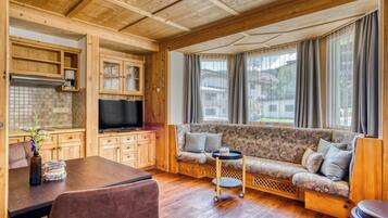 Una habitación acogedora de estilo cabaña con techo de madera, un sofá con cojines, un televisor, una mesa de comedor con sillas y una ventana con cortinas.