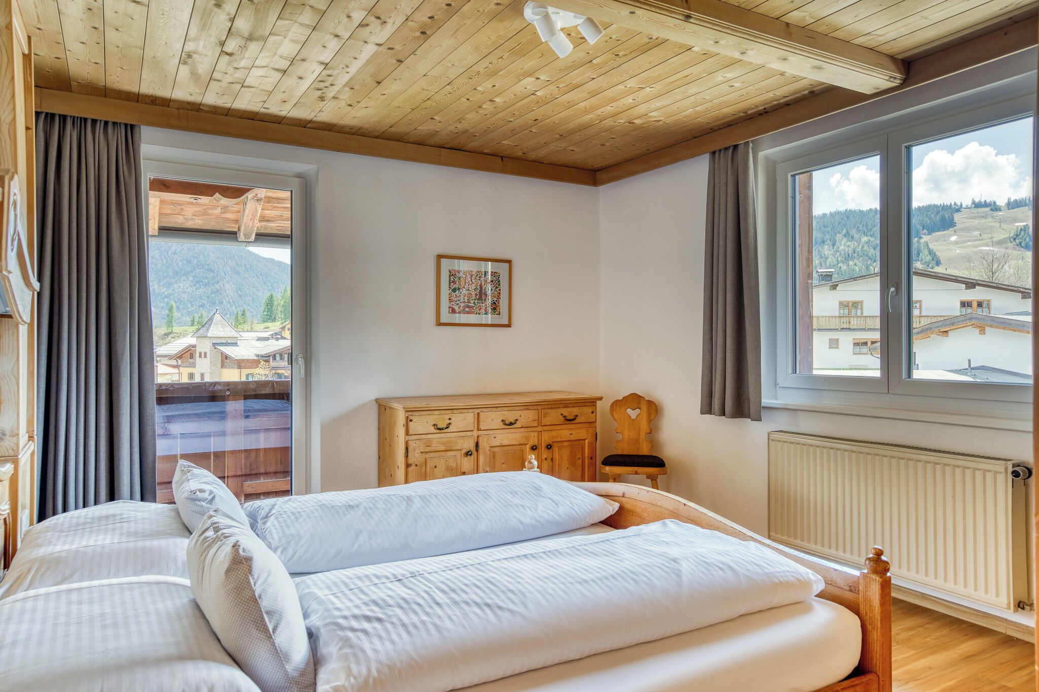 Een slaapkamer met een houten plafond, een groot raam met uitzicht op gebouwen en bergen, een houten dressoir en een bed met witte beddengoed.