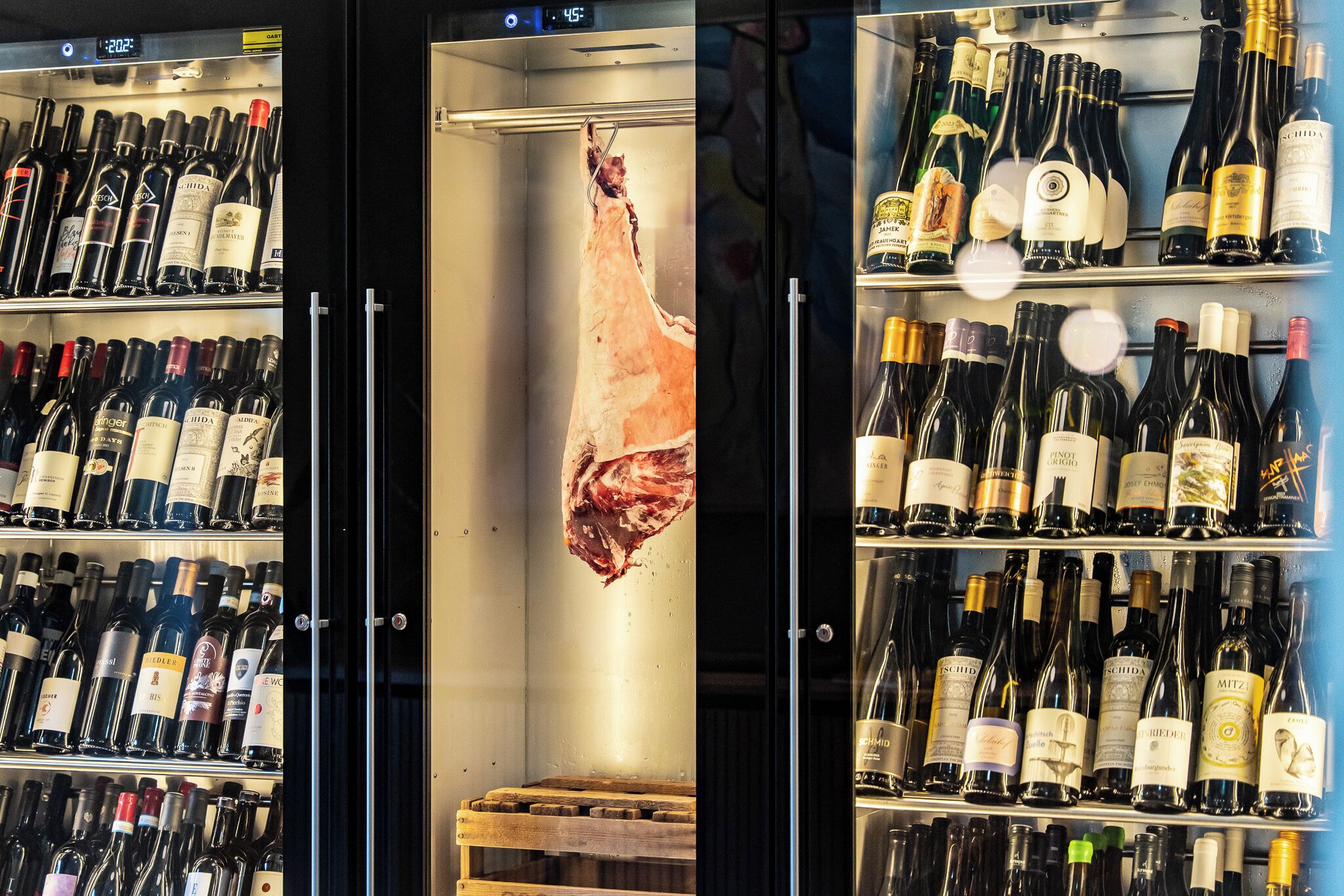 Vitrina refrigerada con varias botellas de vino y un gran trozo de carne colgando del centro.