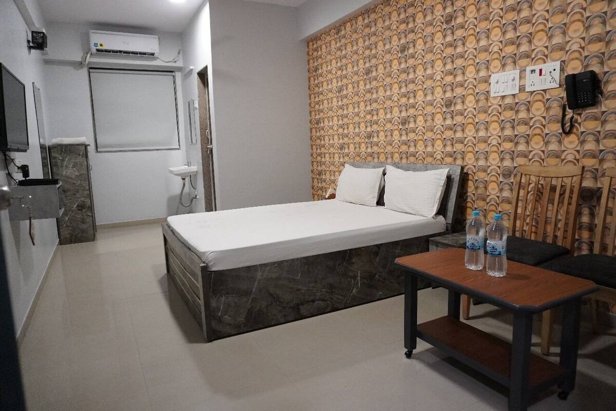 A hotel room with a bed, a small table with water bottles, a chair, a wall-mounted phone, and a wall-mounted air conditioner.