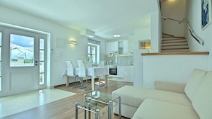 Een moderne woonkamer met een bank, een glazen salontafel en een eethoek met een tafel en stoelen. Er is een keuken met witte kasten en een trap die naar een hoger gelegen verdieping leidt.