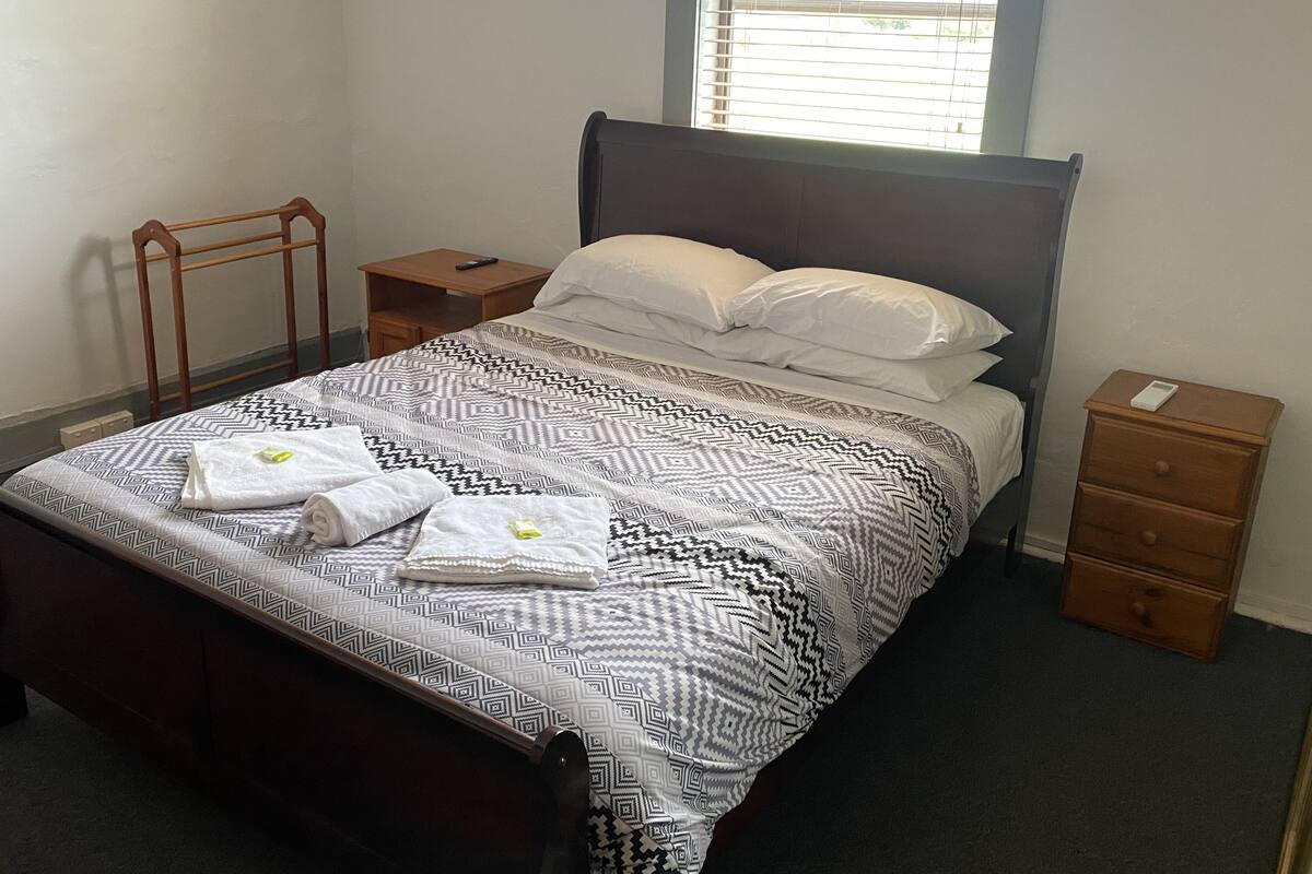 A neatly made bed with a patterned bedspread, two pillows, and a set of towels. There is a wooden nightstand with a remote control beside the bed. A window with blinds is visible in the background.