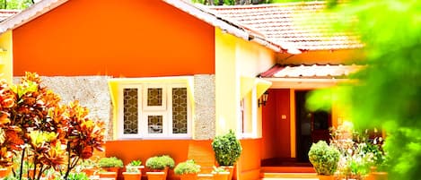 A house with a red roof and orange walls, surrounded by potted plants and a stone pathway.